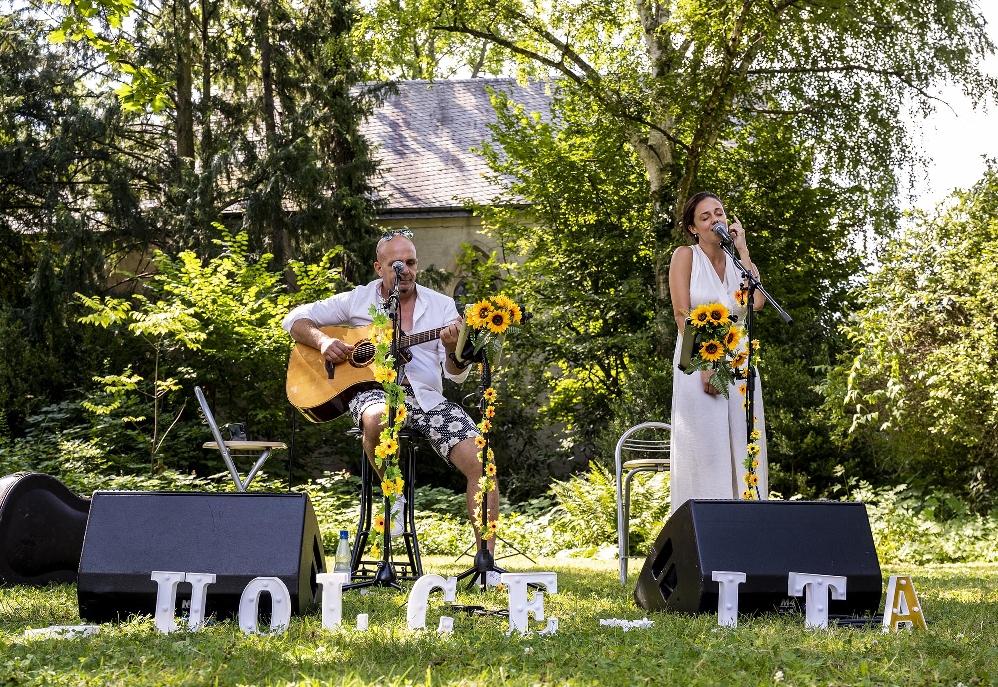 Duo Dolce Vita Acustic alias Vera Della Scala (Gesang) und Raffaele Savoia (Gitarre) spielen in weißer Kleidung und mit Sonnenblumen geschmückt auf der Wiese vor der Adenauerkapelle